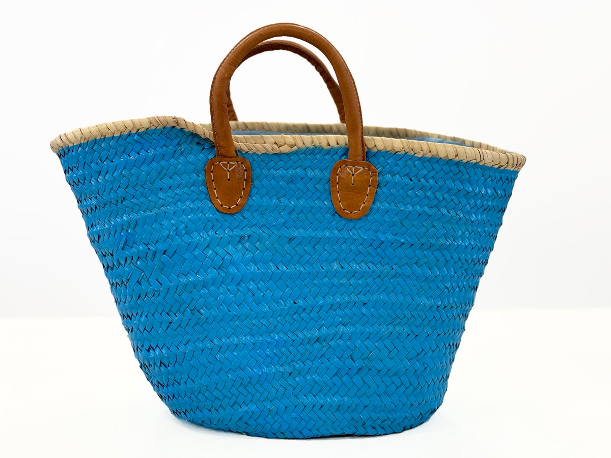 Backside view of turquoise handmade palm leaf basket with natural rim, brown handles and woven texture on white background.