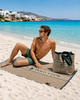 Man sitting on a beach with a branded bag next to him, with a scenic background, unisex classic Greenwich polo towel in extra large size 90x180cm unisex