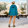 Person wearing a teal jacket with a hood, petrol cooler white sticked framed details around the edges perfect fit with camel brown jeans 