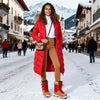 Woman in a red puffer coat and brown boots standing on a snowy street with mountains in the background.