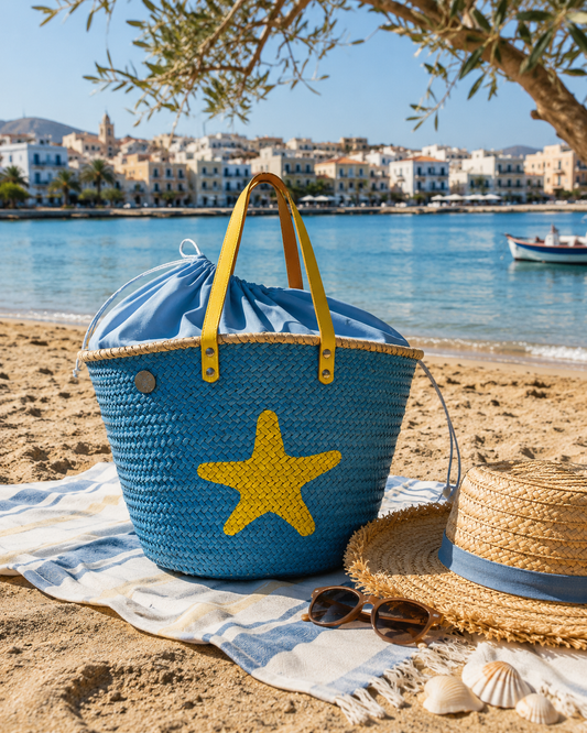 Blue woven bag with a yellow starfish on a sandy beach with ocean and buildings in the background.