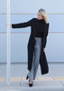 Woman wearing a black coat and gray pants against a light blue wall.
