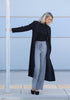 Person wearing a long black coat, black top, and gray pants against a light blue wall.