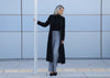 Person wearing a black coat and gray pants standing against a light blue wall.