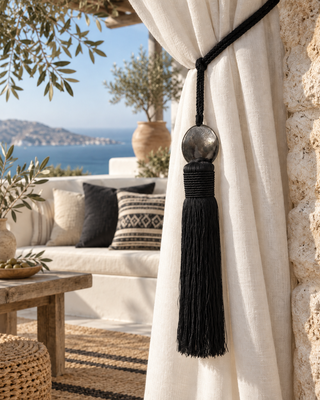 Black Moroccan-style curtain tassel tieback with aluminium disc and satin fringes on a white outdoor curtain in a Cretan Mediterranean setting.