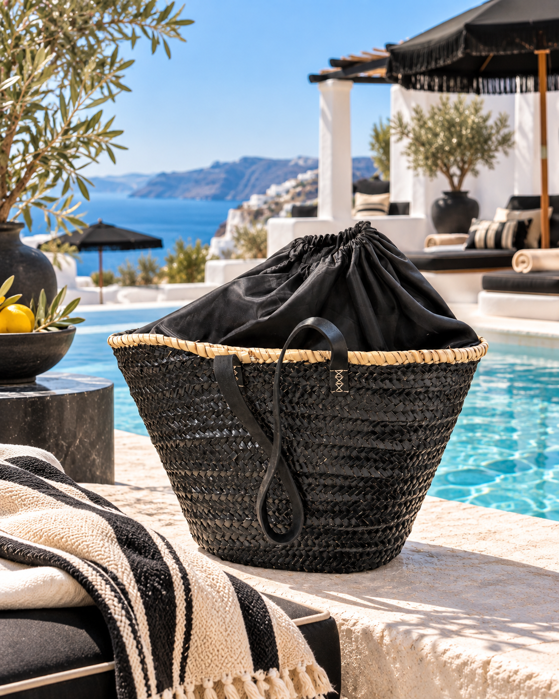 Black raffia beach basket with secure drawstring lining styled at a Mediterranean beach club in Greece.
