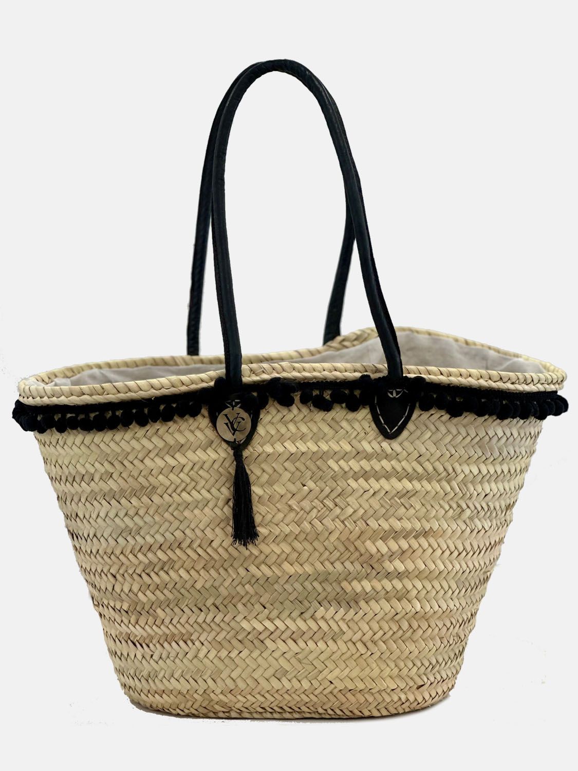 A handmade wicker basket with black leather handles and decorative pom-poms, featuring a textile fall out protection inlet.