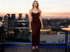 Front view of a woman wearing a brown bodycon slim-fit dress with spaghetti straps on a terrace at sunset.