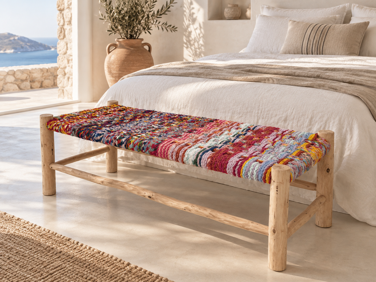 Handmade colourful bench at the foot of a linen bed in a Mediterranean bedroom overlooking the sea.