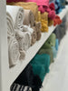 A selection of VVC cotton blankets in various colors neatly folded and displayed on a shelf, with each blanket featuring a pom-pom detail.