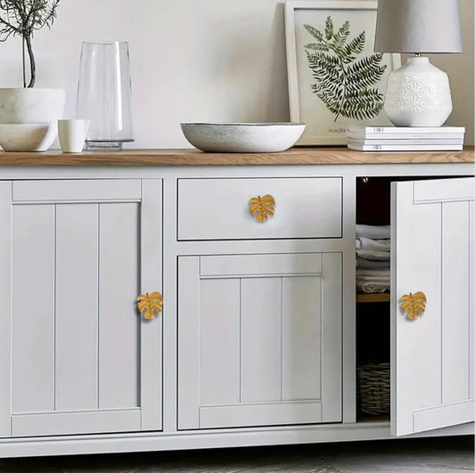 Gold Monstera leaf furniture knobs fitted to a white cabinet in a bright interior.
