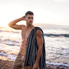 Man by the sea with a Greenwich Polo Club 4077 black and beige striped beach towel draped over his shoulder.