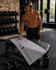 Tattooed male model in a gym picking up a gray Greenwich Polo Club microfiber gym towel 4184 from a workout bench, lifestyle fitness image for VVC SHOP Sitia, Crete.
