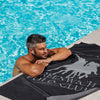 Male model relaxing beside a pool with the Greenwich Polo Club black microfiber beach towel 5214 in Crete.