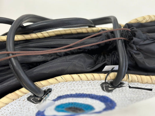 Close-up of large white sequin evil eye basket with black handles, straw rim and blue evil eye design.