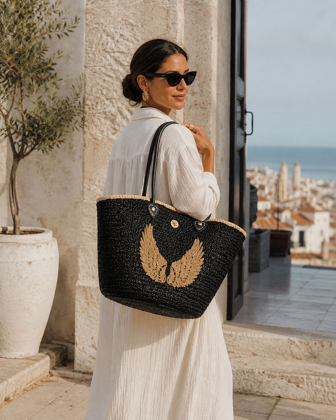 Luxury VVC black sequin beach basket with gold angel wings detail carried by a stylish woman in Sitia, Crete. Handmade oversized summer tote with leather handles, Mediterranean resort fashion styling and elegant coastal lifestyle inspiration for beach, travel and shopping.