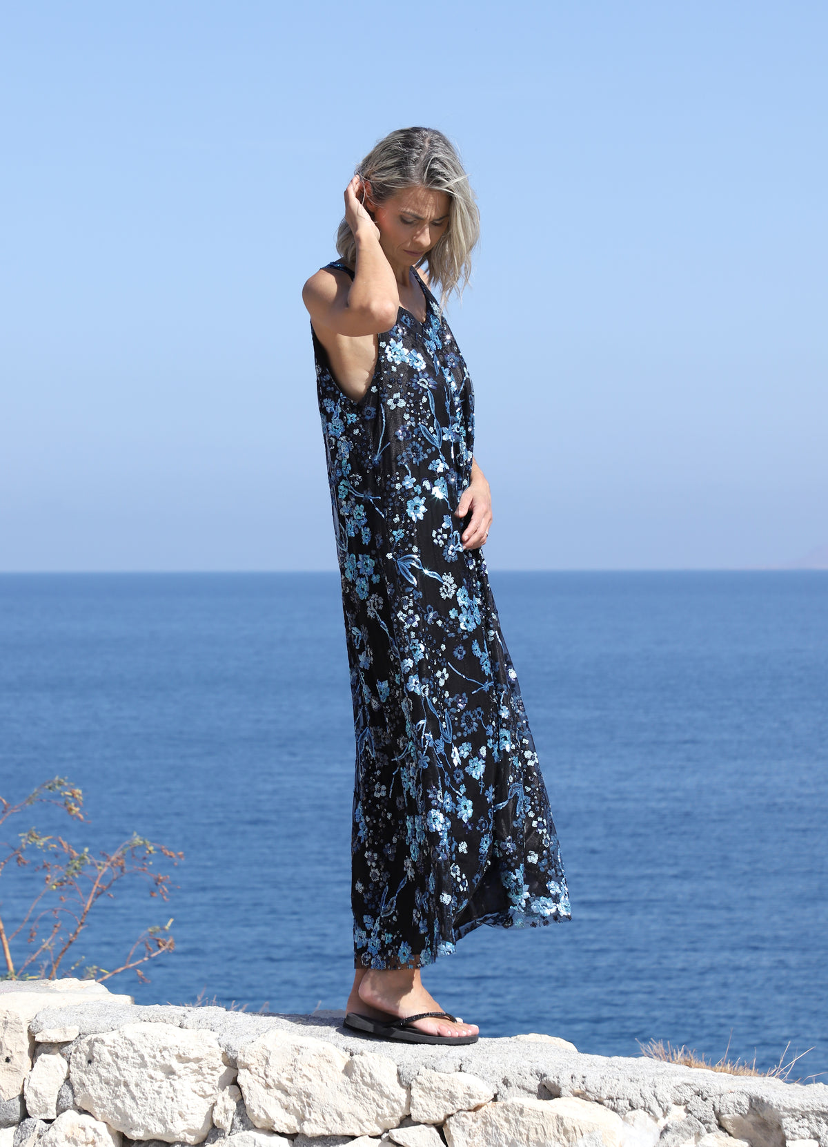 Front-side view of a woman wearing the VVC blue-and-black floral sequin mullet dress on a rocky coastline.