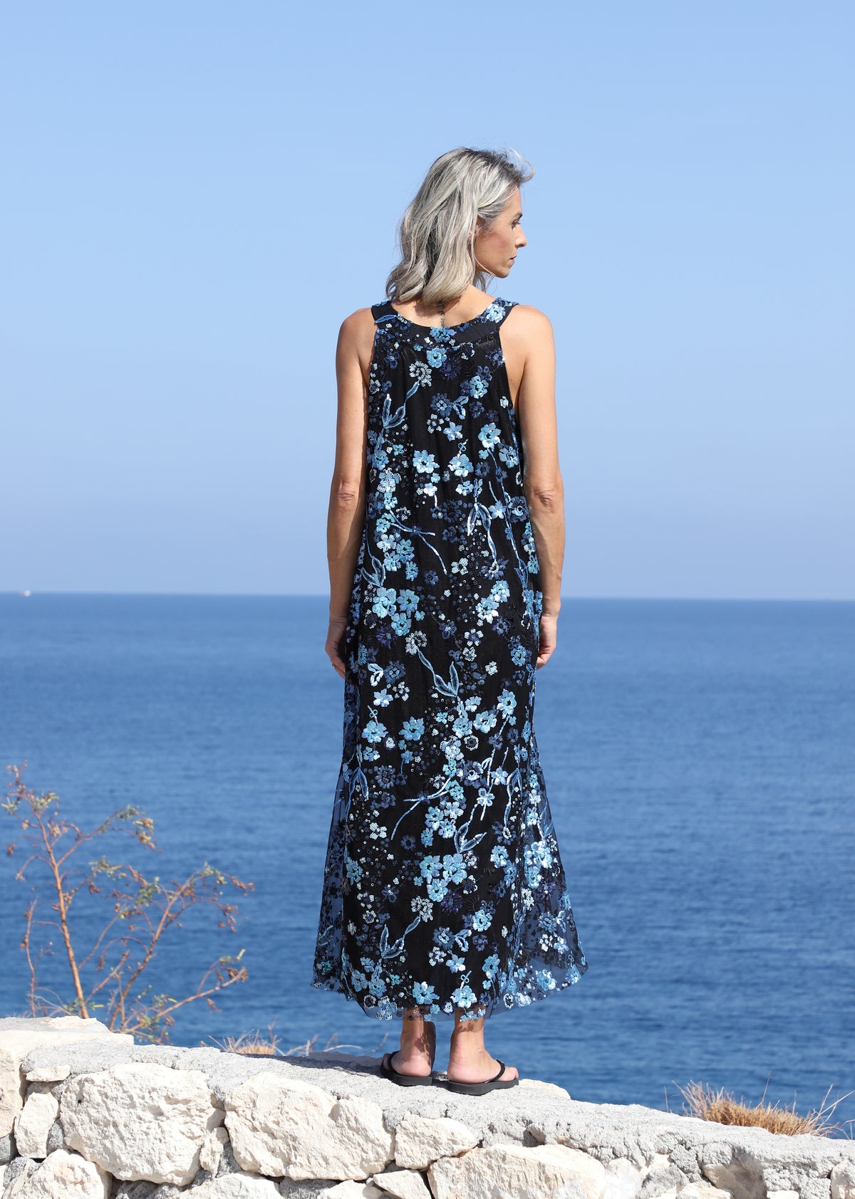 Full-length front view of the VVC blue-and-black floral sequin mullet evening dress beside the sea.