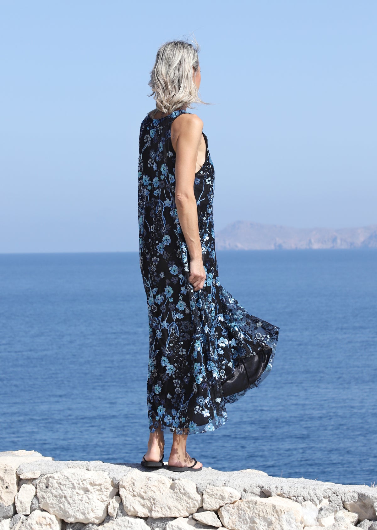 Side view of the VVC blue-and-black floral sequin mullet dress beside the sea, showing its long asymmetric back hem.