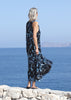 Side view of the VVC blue-and-black floral sequin mullet dress beside the sea, showing its long asymmetric back hem.