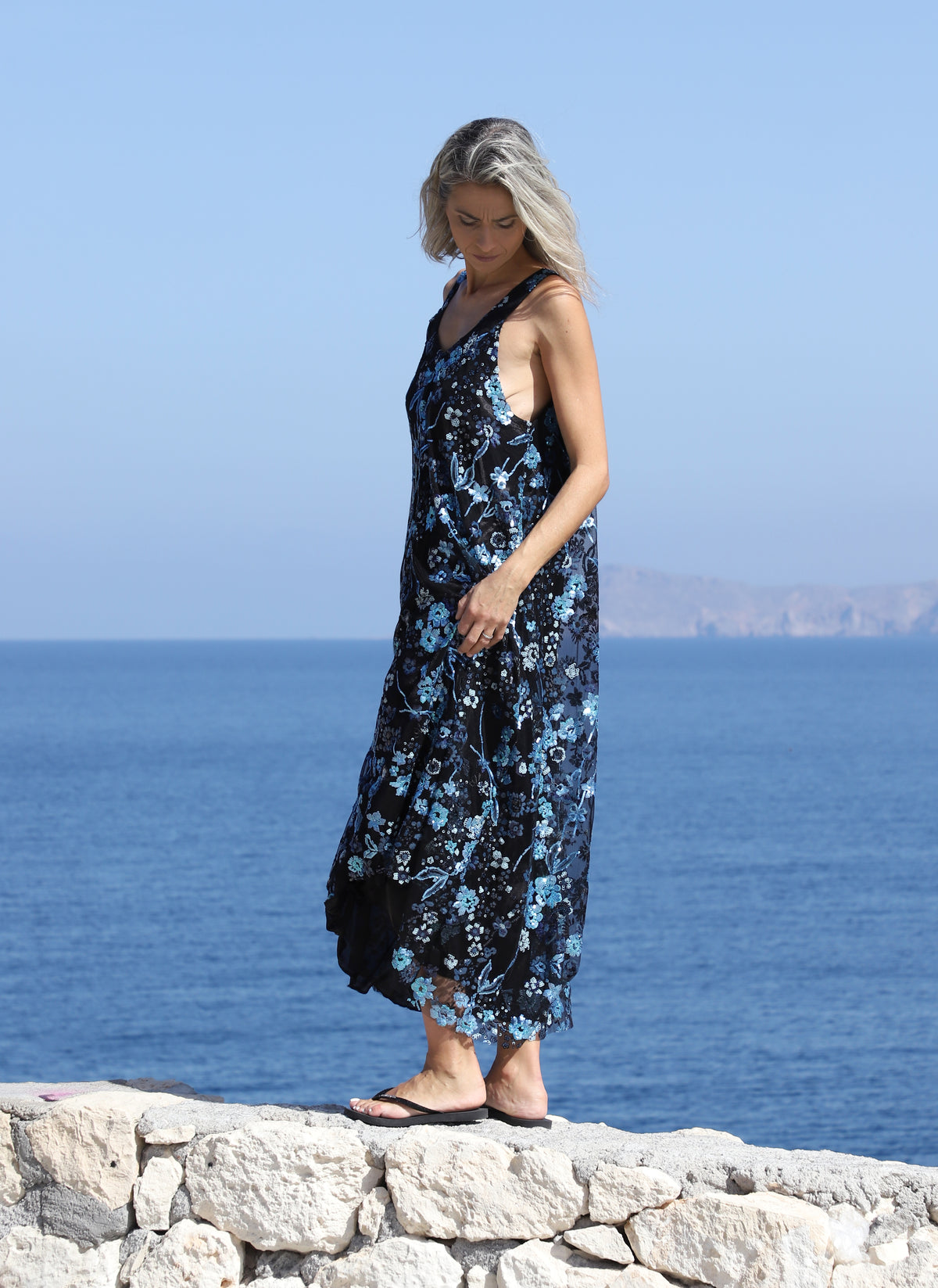 Woman walking beside the sea in the VVC blue-and-black floral sequin mullet dress, showing the movement of its flowing hem.
