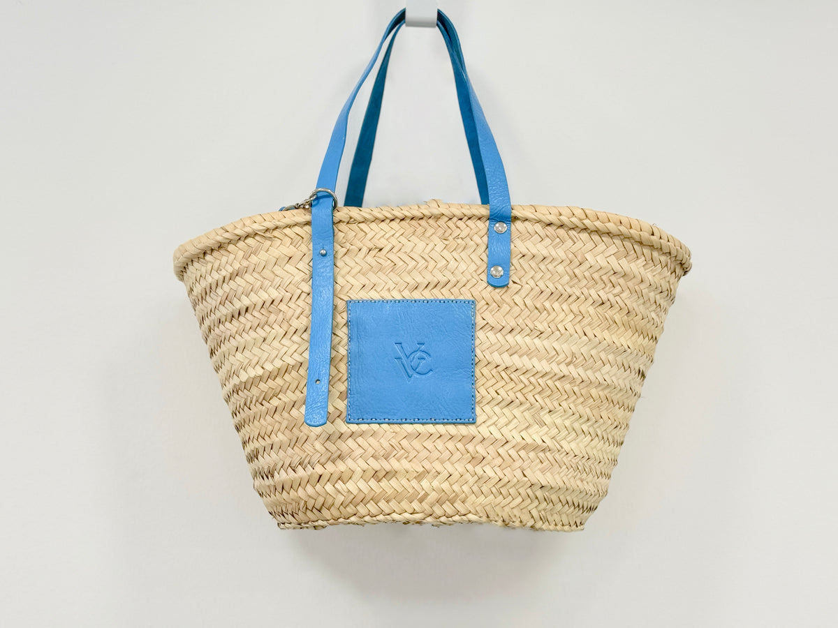 Side view of handmade VVC™ woven palm leaf basket with blue leather details and shoulder strap. Practical shopping tote and resort-style travel bag from Sitia, Crete.