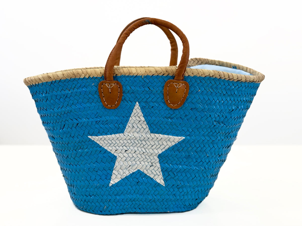 A handmade palm leaf basket painted turquoise with a white star on one side, featuring a brown short handle.