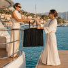 Two women passing an oversized VVC™ black satin fringe raffia basket across a private yacht railing during a luxury Mediterranean holiday.