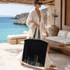 Woman carrying a large VVC raffia basket covered in long black satin fringes at a luxury beach club in Crete.