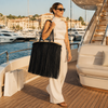 Tattooed model carrying an oversized VVC black satin fringe raffia basket over her shoulder during luxury superyacht shopping in the Mediterranean.