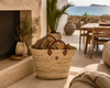 Large handmade palm leaf basket with camel brown leather handles used for stylish firewood storage in a luxury Mediterranean living space.