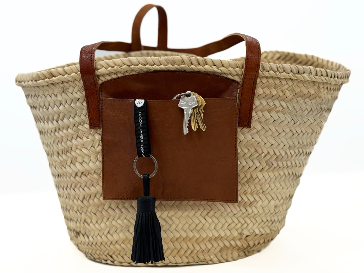 Front view of a handmade VVC™ palm leaf basket with folded leather handles, leather mobile phone pocket and artisan craftsmanship, perfect as a shopping basket, beach tote or home décor accessory in Crete.
