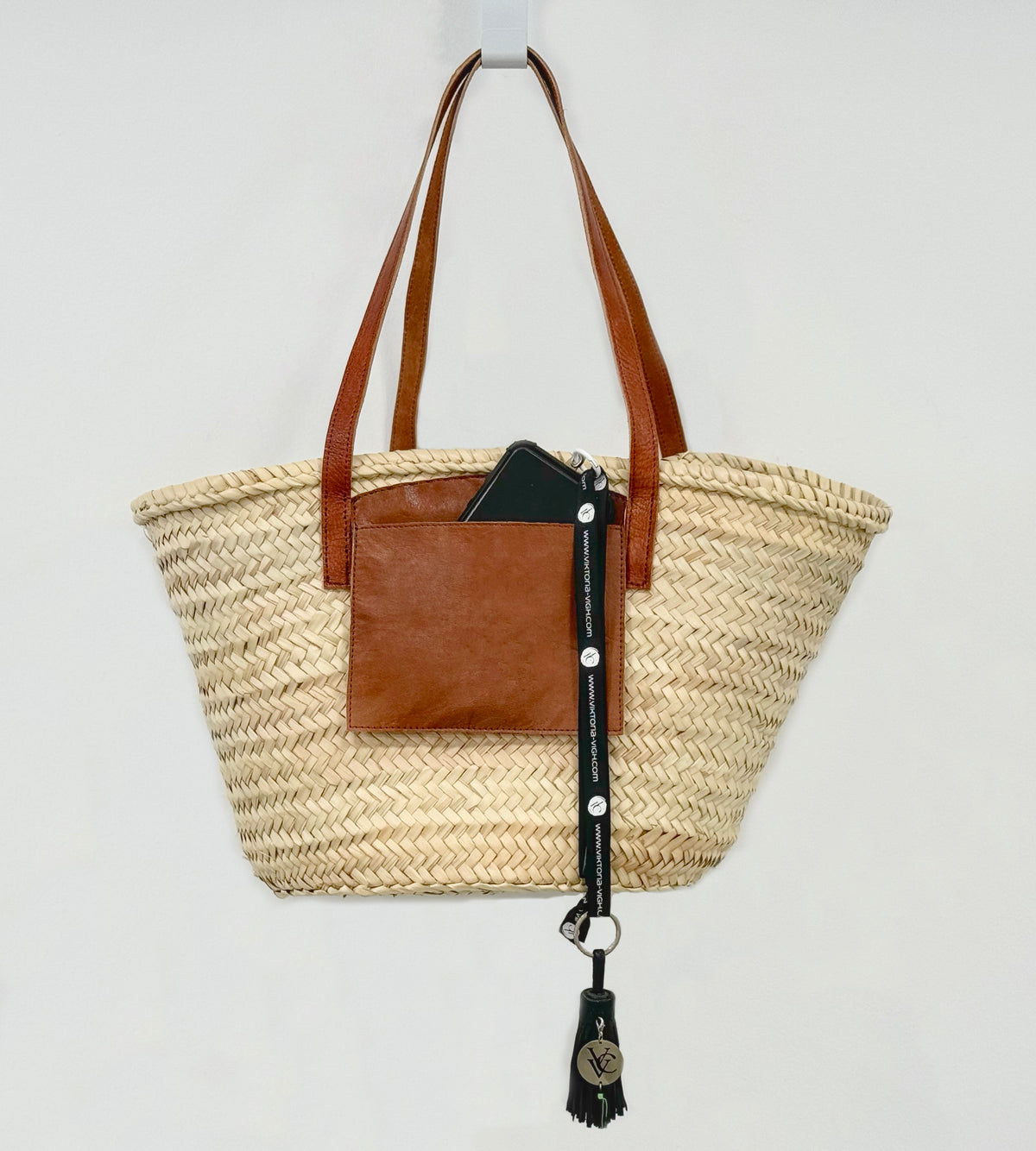 VVC™ handmade palm leaf basket with long brown leather handles and practical leather pocket, displayed hanging as a stylish Mediterranean storage, shopping and travel basket inspired by life in Crete, Greece.