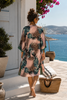 Rear three-quarter walking view of the VVC pink silk cardigan, showing the flowing back, green lurex cuffs and side slits on a Greek island terrace.