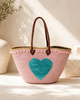 Handmade VVC beach basket covered in soft pink sequins with a large turquoise heart applique, baby pink pompom detail and brown leather handles. Artisan-crafted boho luxury basket with up to 100 litres capacity, available at VVC Shop Sitia and online at viktoria-vigh.com.
