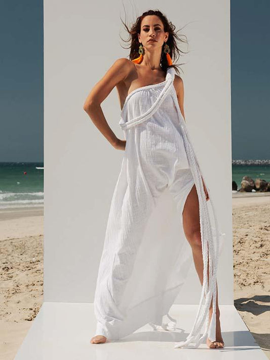 Woman wearing the VVC white one-shoulder cotton maxi dress with macramé shoulder detail on a Mediterranean beach