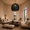 XXXL black handmade bast-fibre pendant lampshade, 105 × 95 cm, casting woven light patterns in a Greek island living room with arched windows, an open door and sea views, styled for VVC SHOP Sitia, Crete.