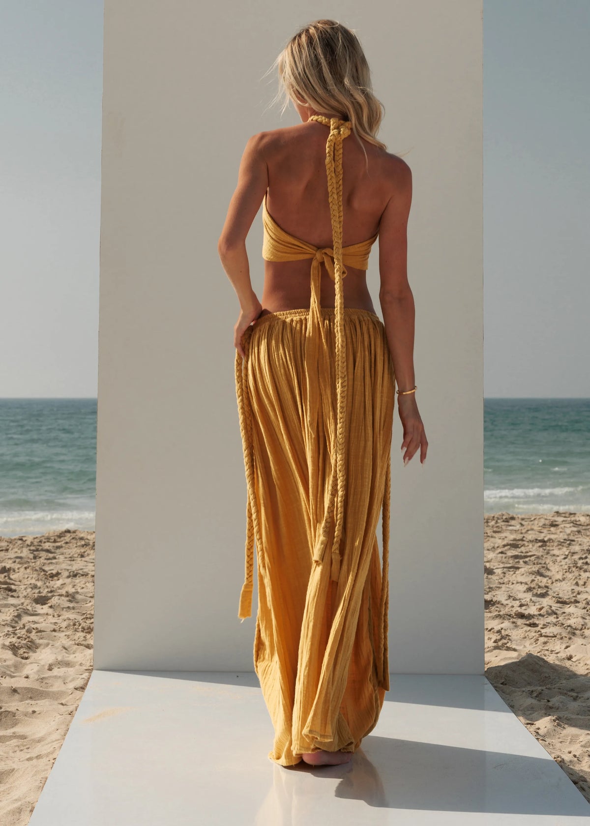 Full-length back view of the ZOE Zena mustard-yellow set showing its braided halter ties and flowing low-waist maxi skirt.