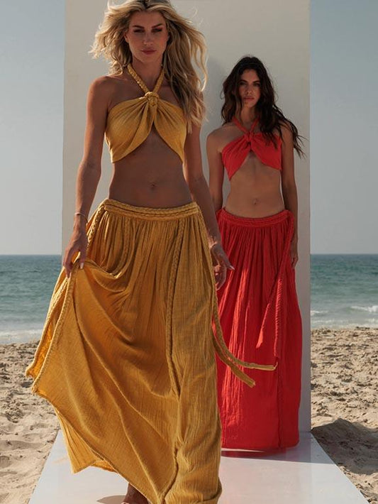 Two models wearing halter neck crop tops and matching skirts in yellow and orange, standing on a beach with the ocean in the background.